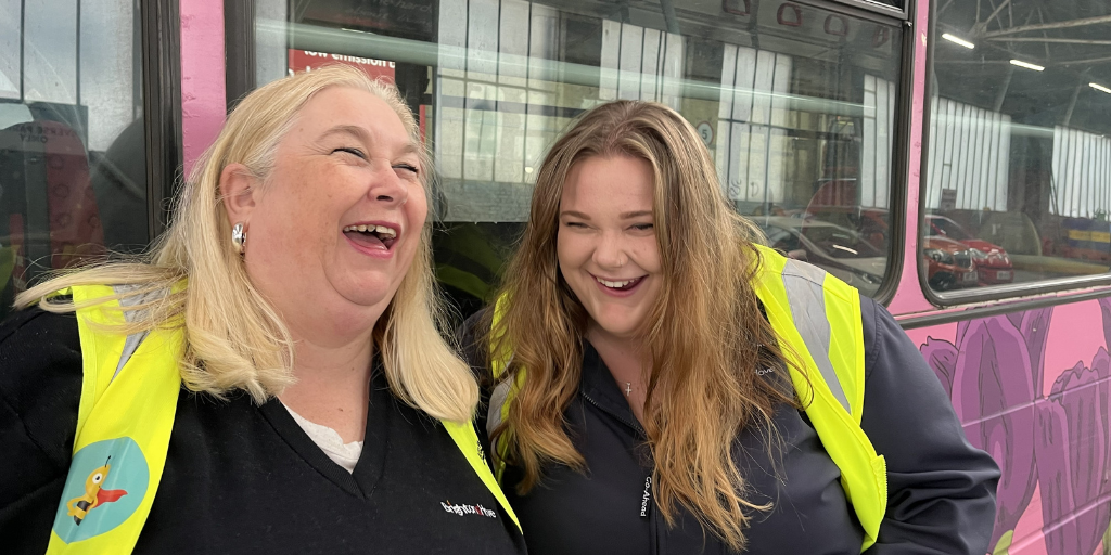 two women wearing hi-vis jackets laughing in front of a bus