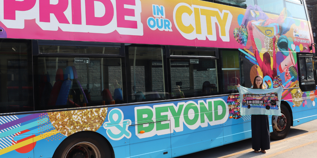 lucy standing in front of the pride bus holding a crochet blanket of the bus